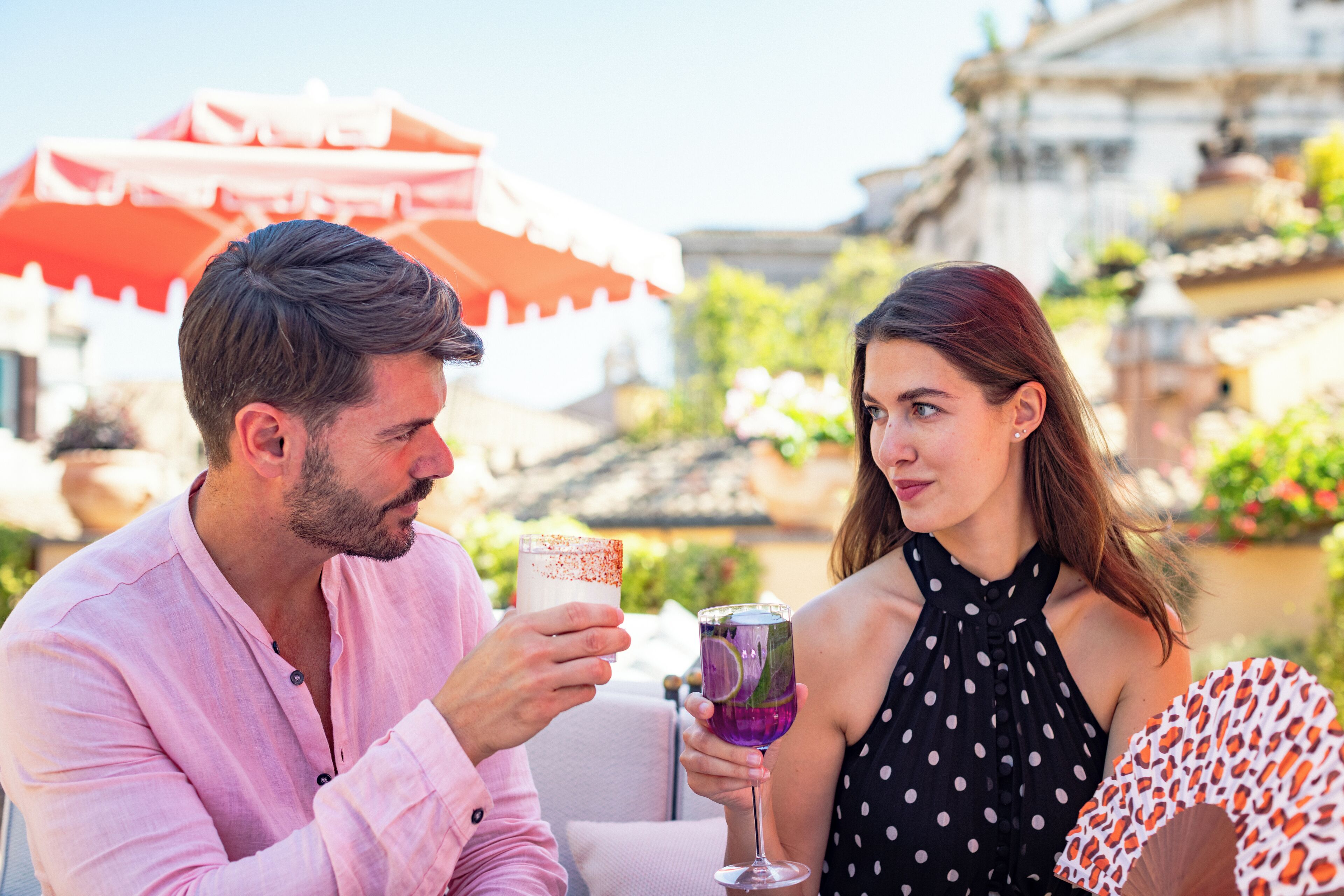 A couple sitting at an outdoor table with drinks, a red umbrella, and a building with traditional architecture in the background.