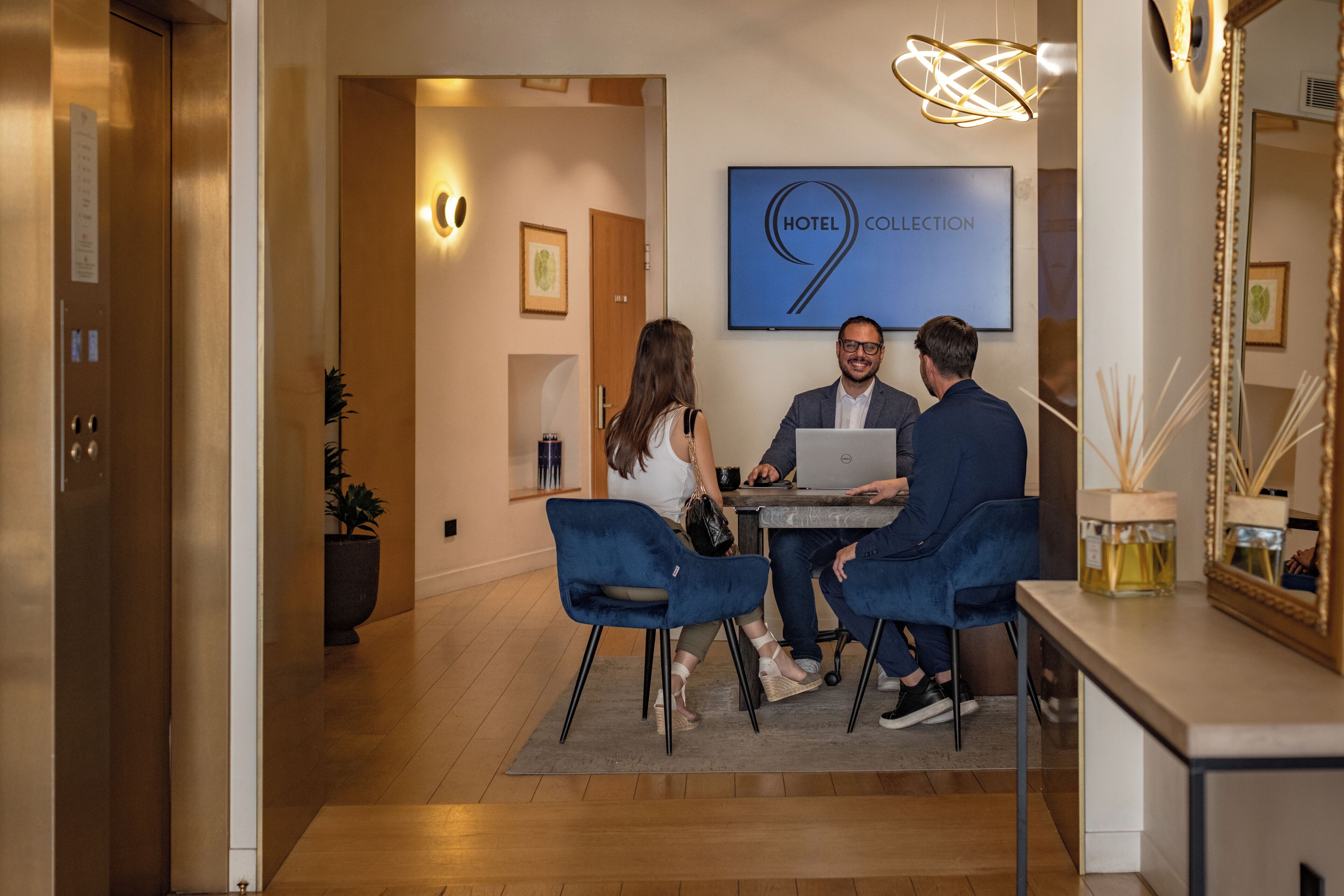 Eine Hotelhalle mit einer Rezeption, einem großen gerahmten Schild mit der Aufschrift "HOTEL COLLECTION" und einem modernen Kronleuchter.