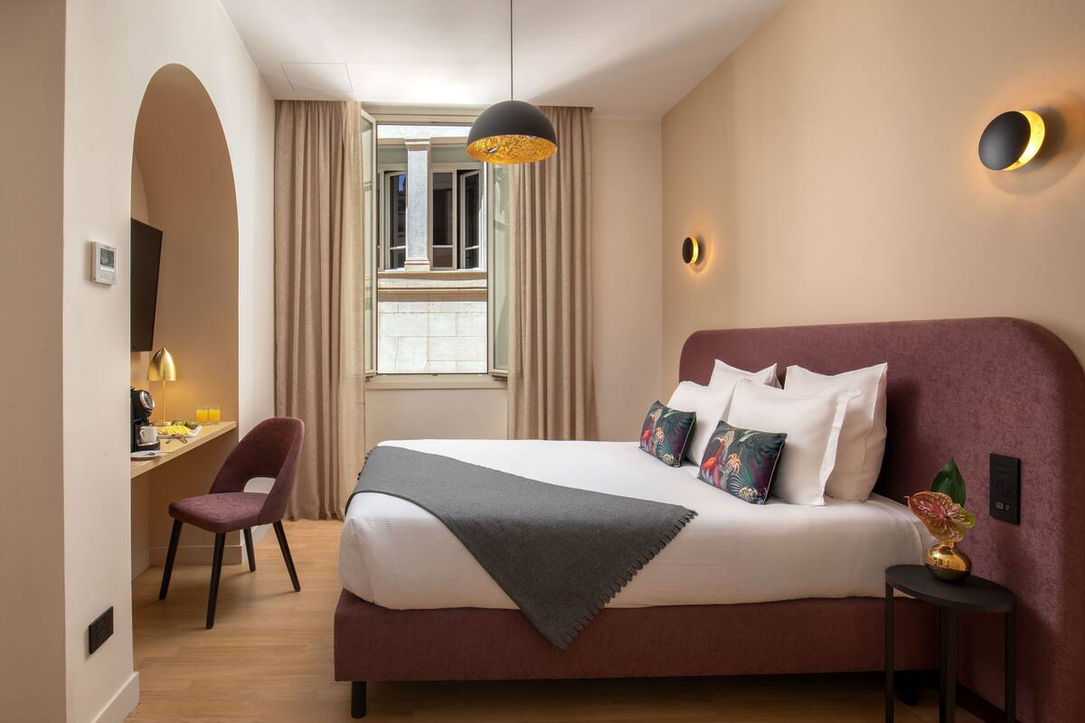 Une chambre d’hôtel avec un grand lit, un bureau avec une chaise, une fenêtre avec des rideaux et un luminaire fixé au mur.