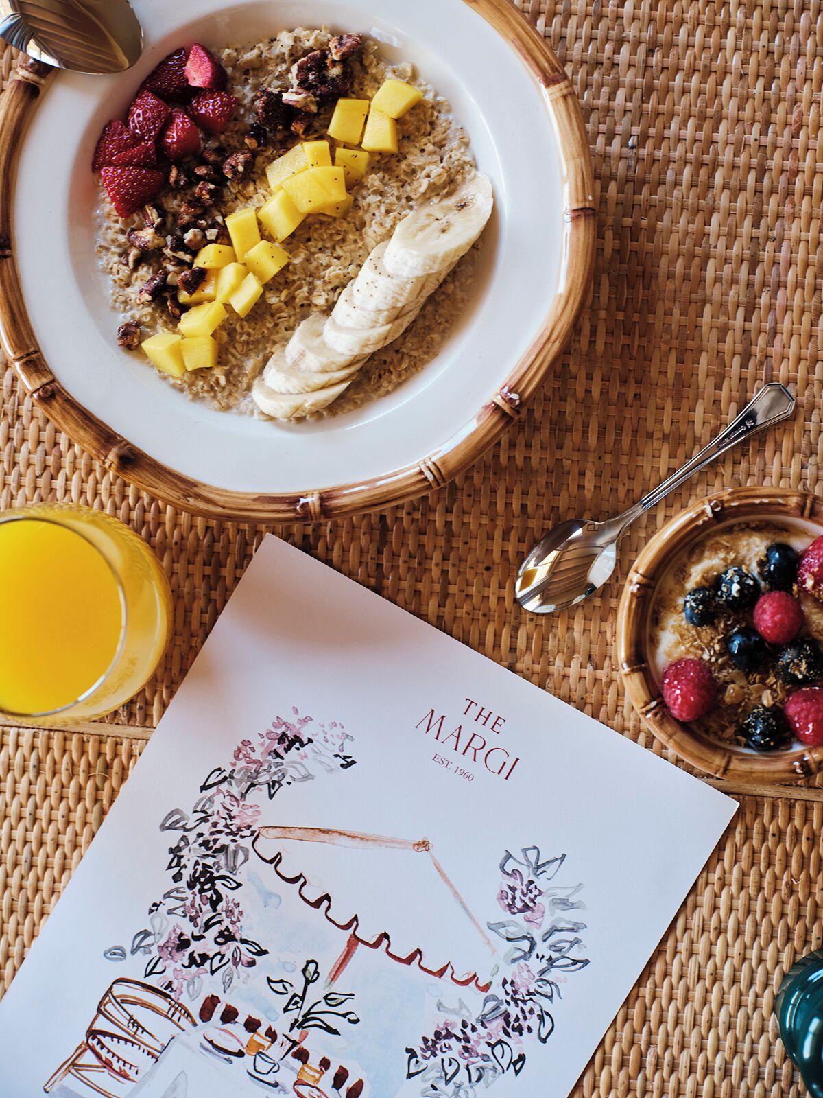 Un desayuno con un tazón de avena cubierto de fresas, mango y rodajas de plátano, un vaso de jugo de naranja y una tarjeta con diseño floral.