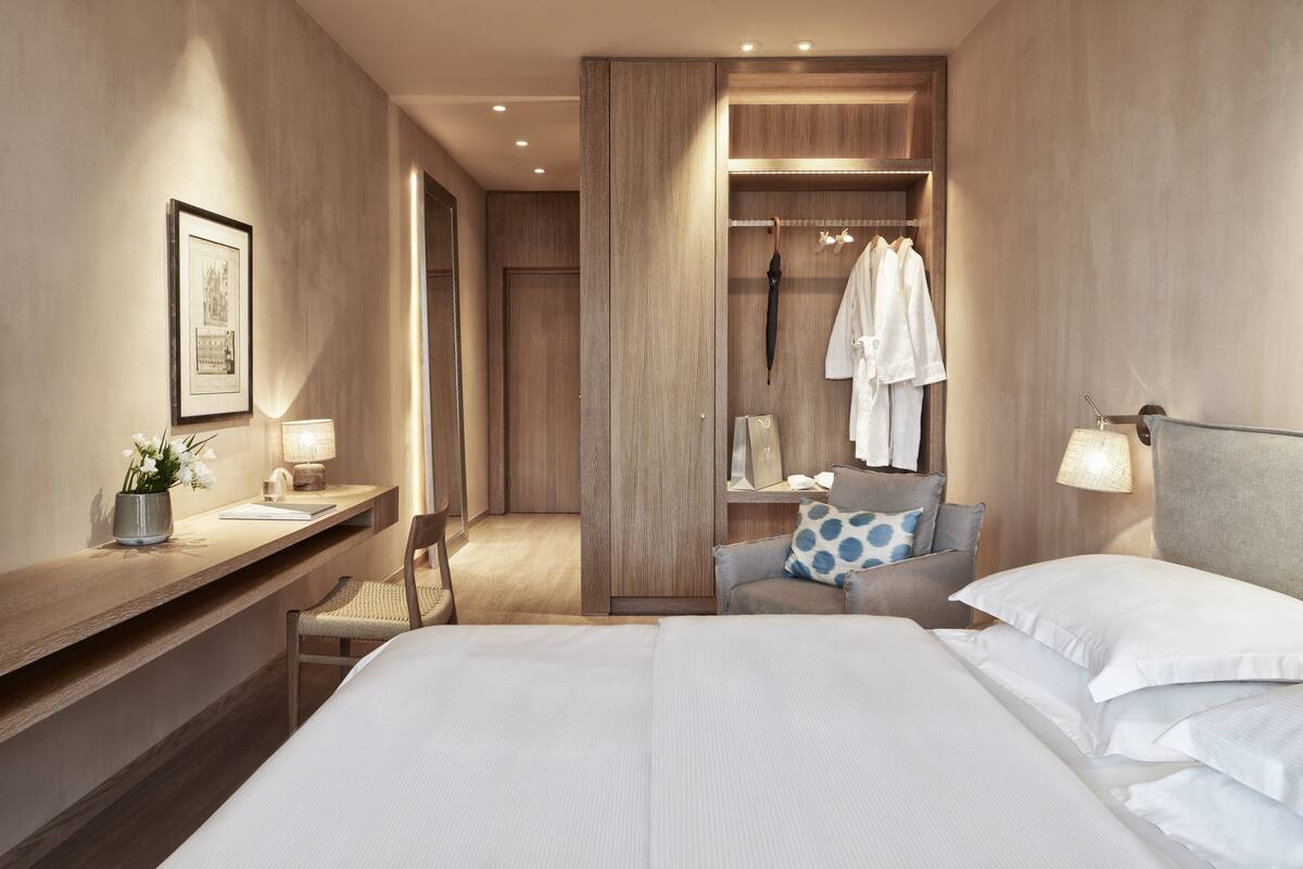 A modern hotel room with a large bed, a wooden wardrobe, a vanity with a mirror, and a seating area with a chair and a small table.