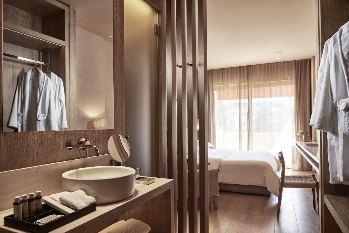 A modern hotel room with a large mirror, a round sink, a bedside table with toiletries, a bed, and a view of the city through large windows.