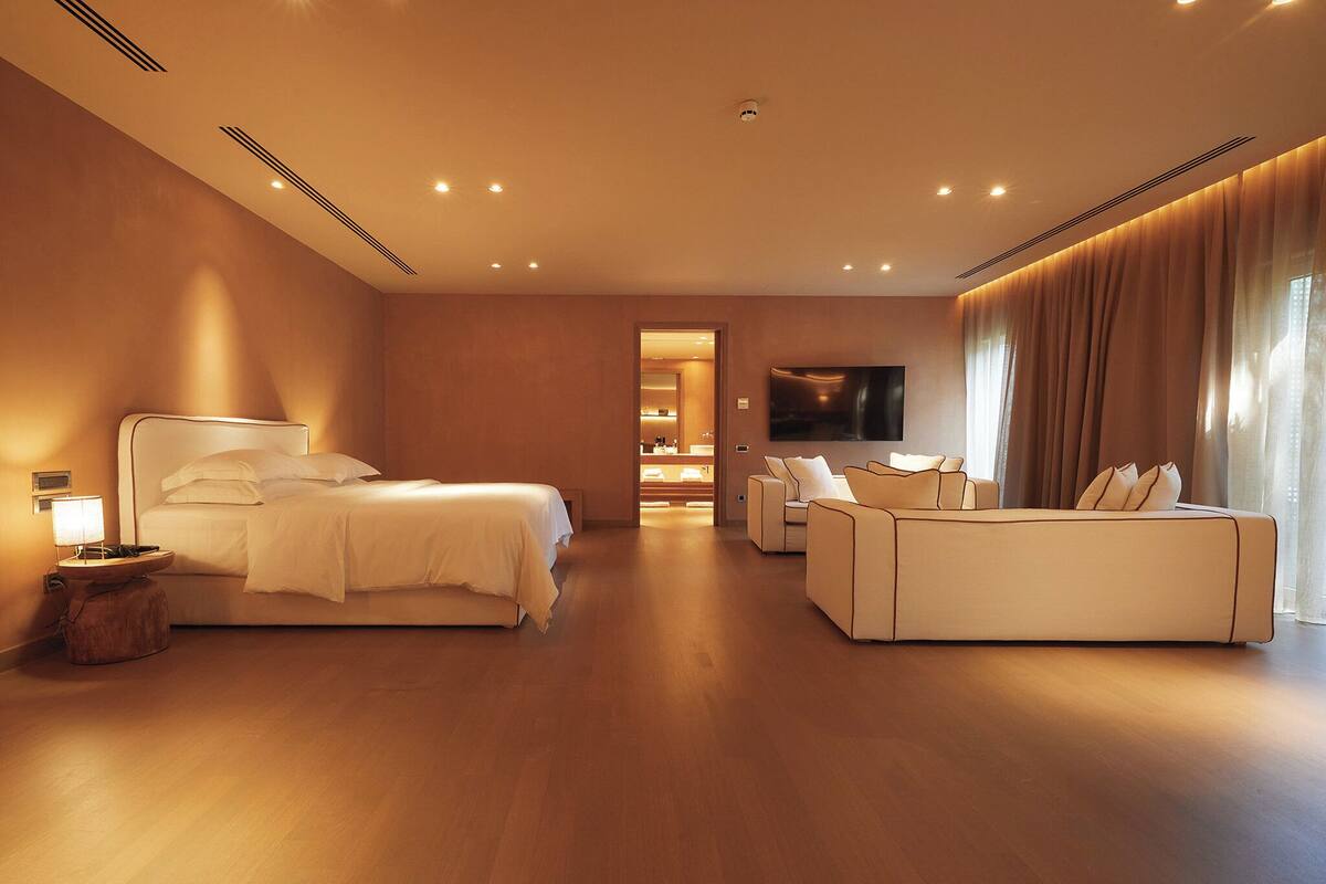 A modern hotel room with a large bed, a sofa, a TV, and a bathroom visible through an open door.