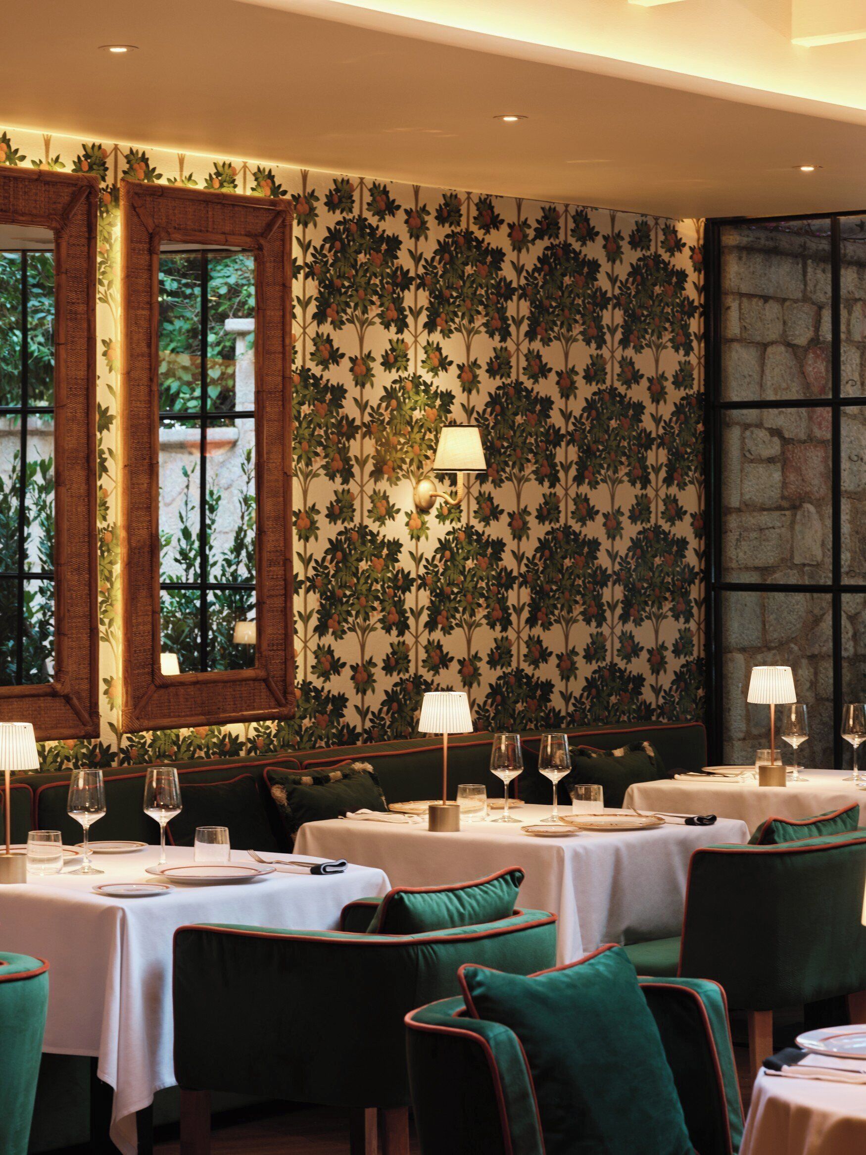 A dining area with tables set for two, featuring green upholstered chairs and a patterned wallpaper.