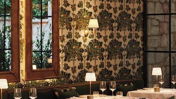 A dining area with tables set for two, featuring green upholstered chairs and a patterned wallpaper.