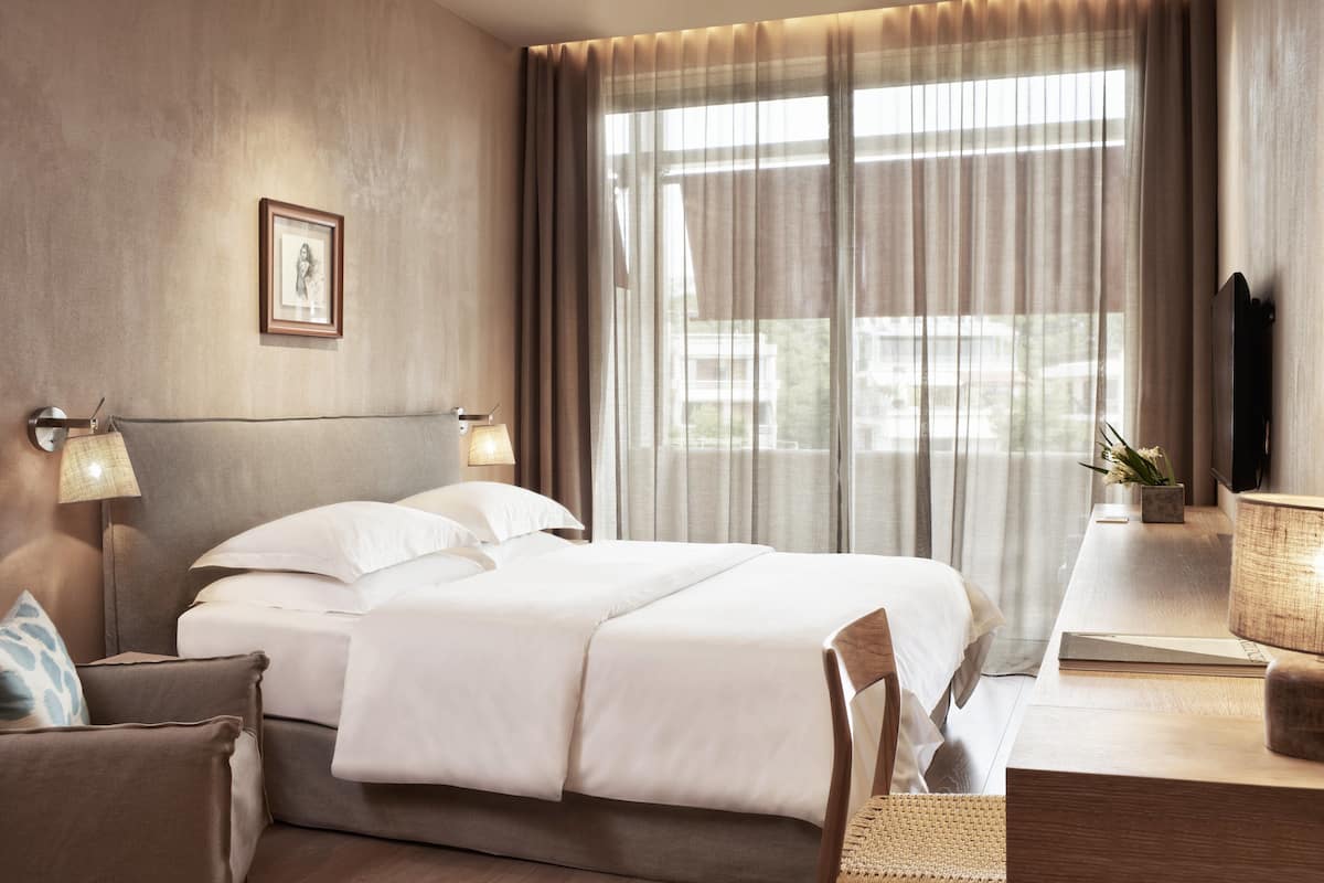 A hotel room with a large bed, a bedside table with a lamp, a chair, and a view of the outdoors through sheer curtains.