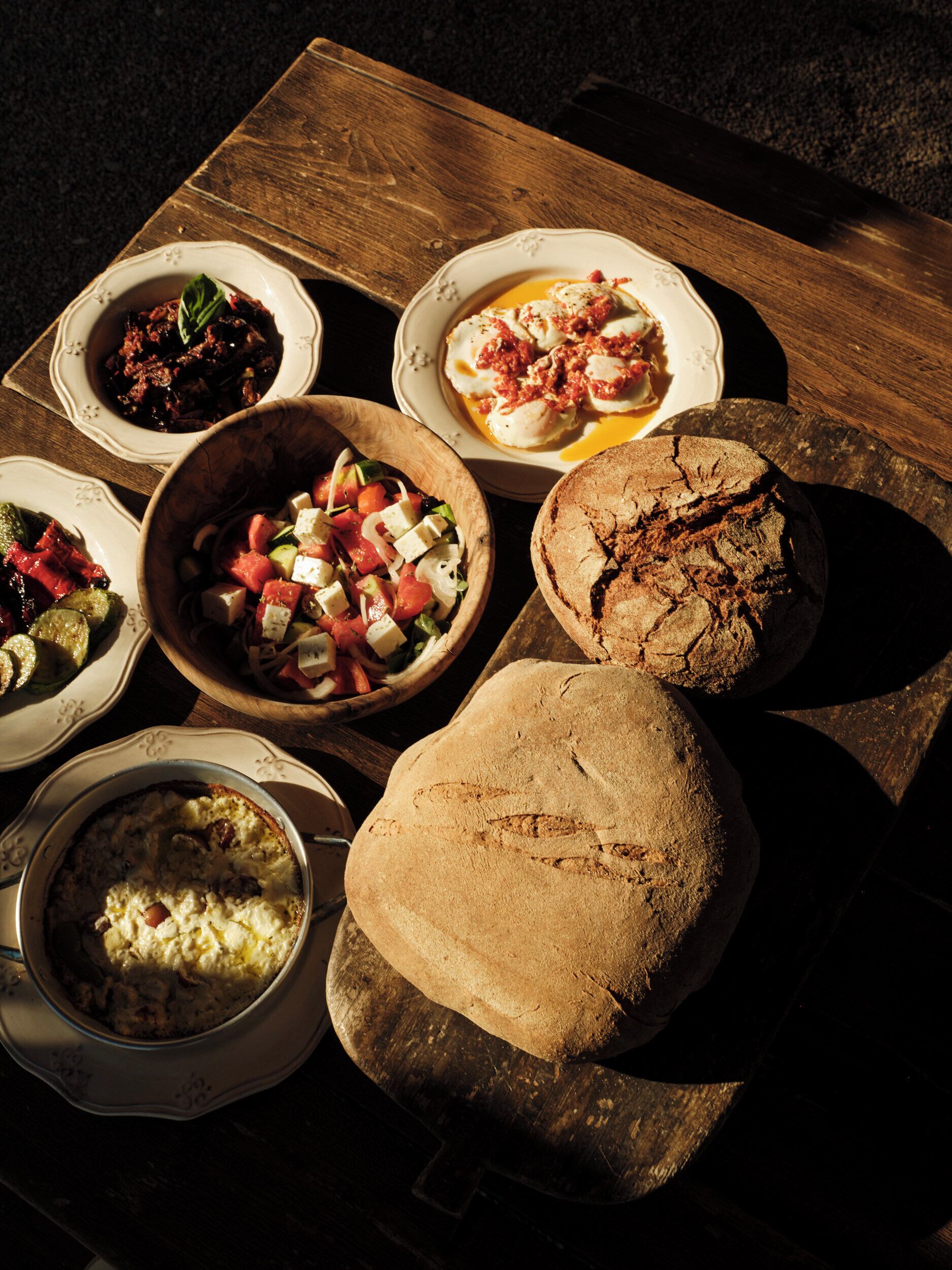 A wooden table with various dishes including a bowl of salad, a plate of eggs, and a loaf of bread.