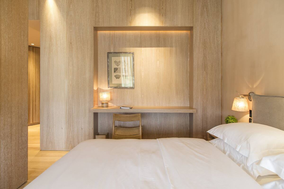 A hotel room with a wooden wall, a desk with a chair, a bed, and a nightstand with a lamp.