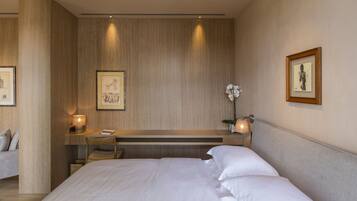 A modern hotel room with a large bed, a desk with a chair, a small table with a lamp, and framed artwork on the walls.