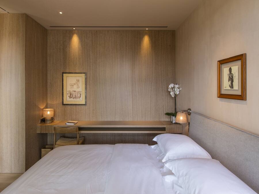 A modern hotel room with a large bed, a desk with a chair, a small table with a lamp, and framed artwork on the walls.