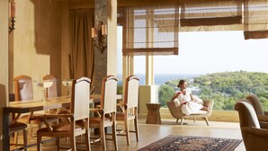 A dining area with a table set for four, a large window with a view of a green landscape, and a patterned rug on the floor. - The Margi (Vari-Voula-Vouliagmeni)