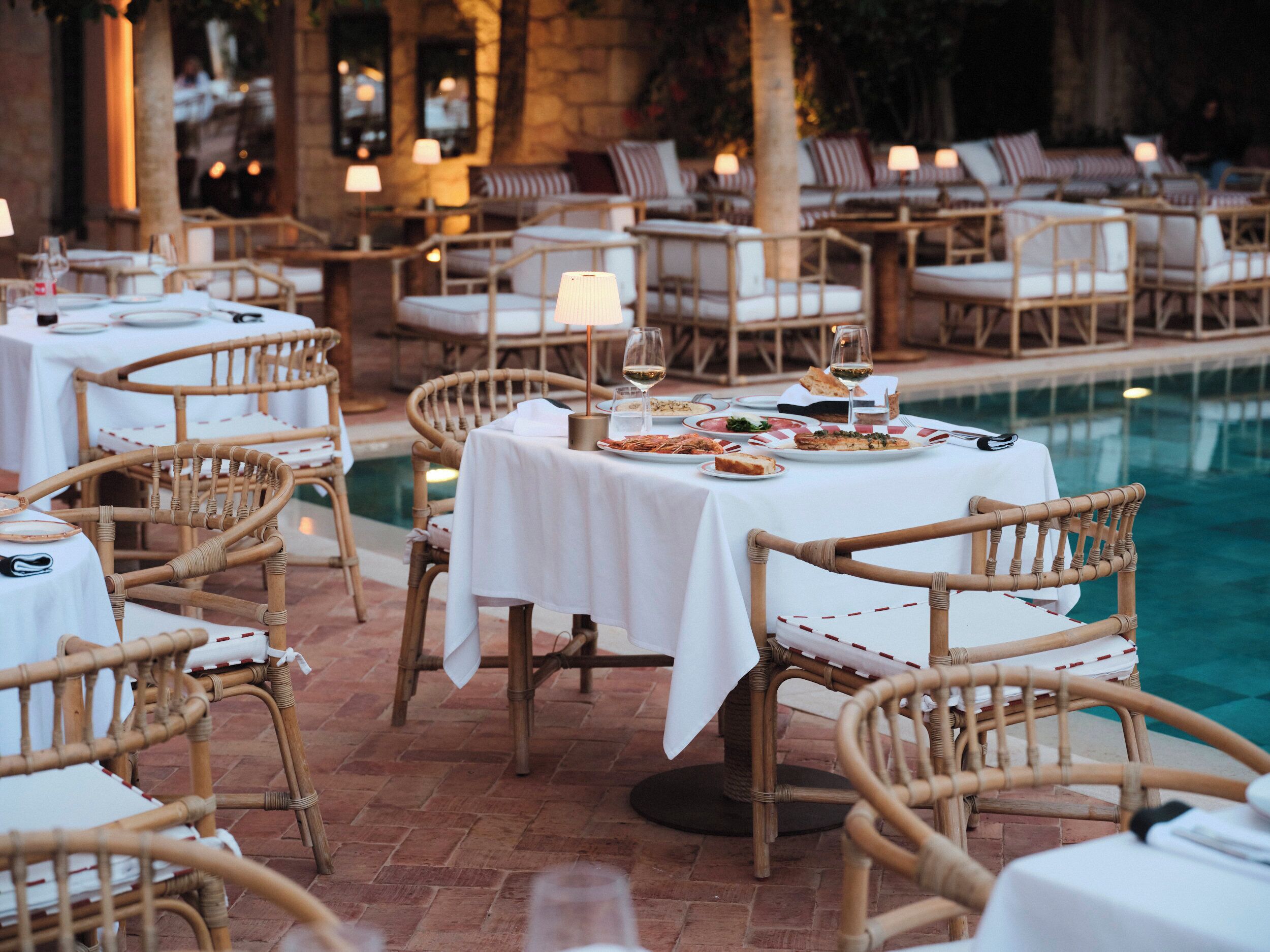 A poolside dining area with wicker chairs and tables, a pool, and a stone wall in the background.