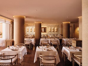 A restaurant with multiple tables and chairs, large columns, and patterned wallpaper. - The Margi (Vari-Voula-Vouliagmeni)