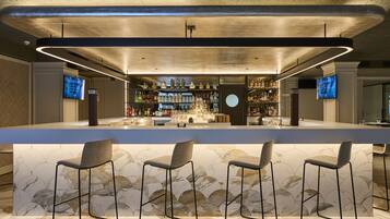A modern bar area with a marble countertop, three bar stools, and a variety of bottles and glasses.