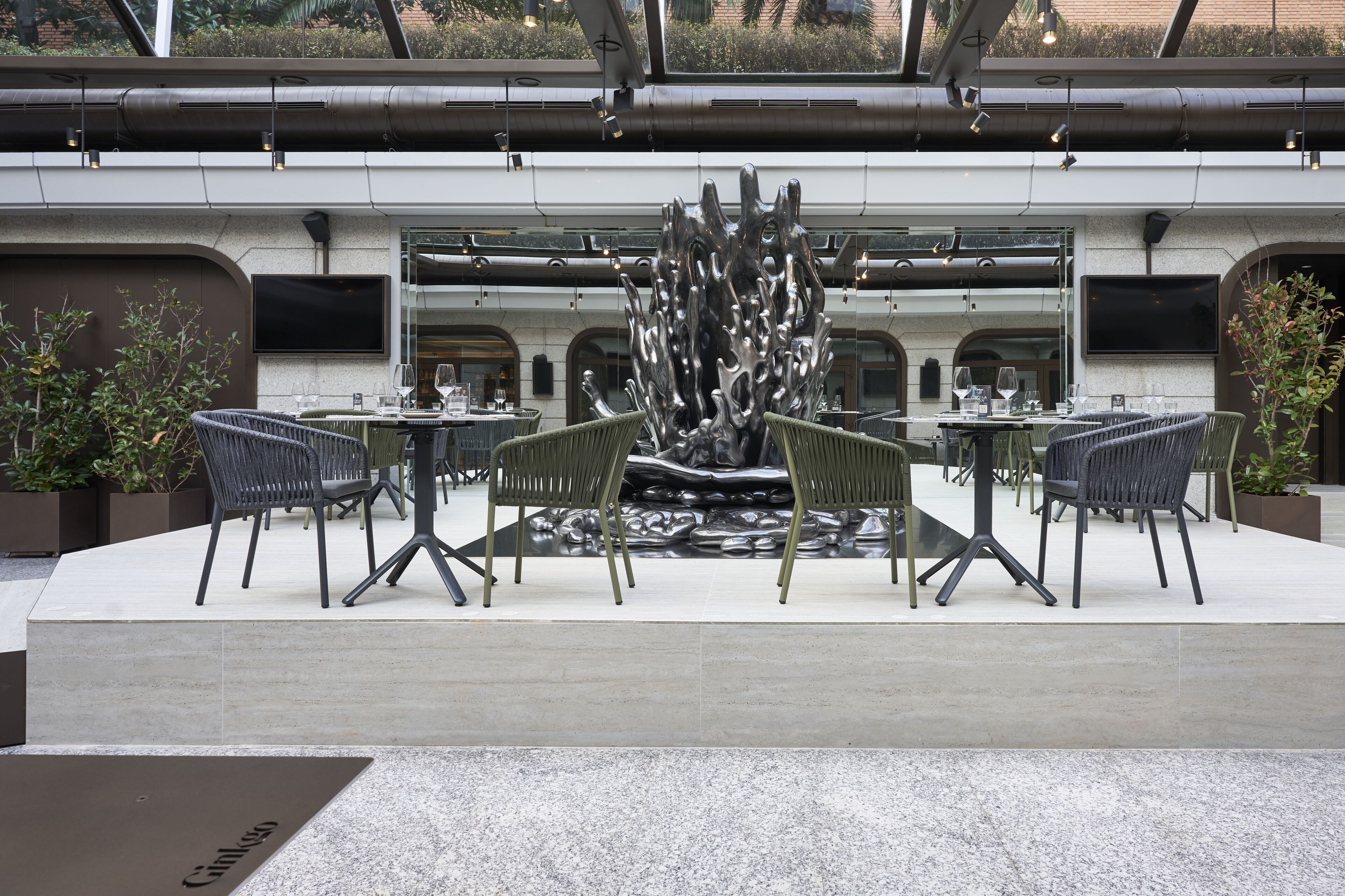 An outdoor dining area with modern chairs and tables, a central sculpture, and a large window in the background.