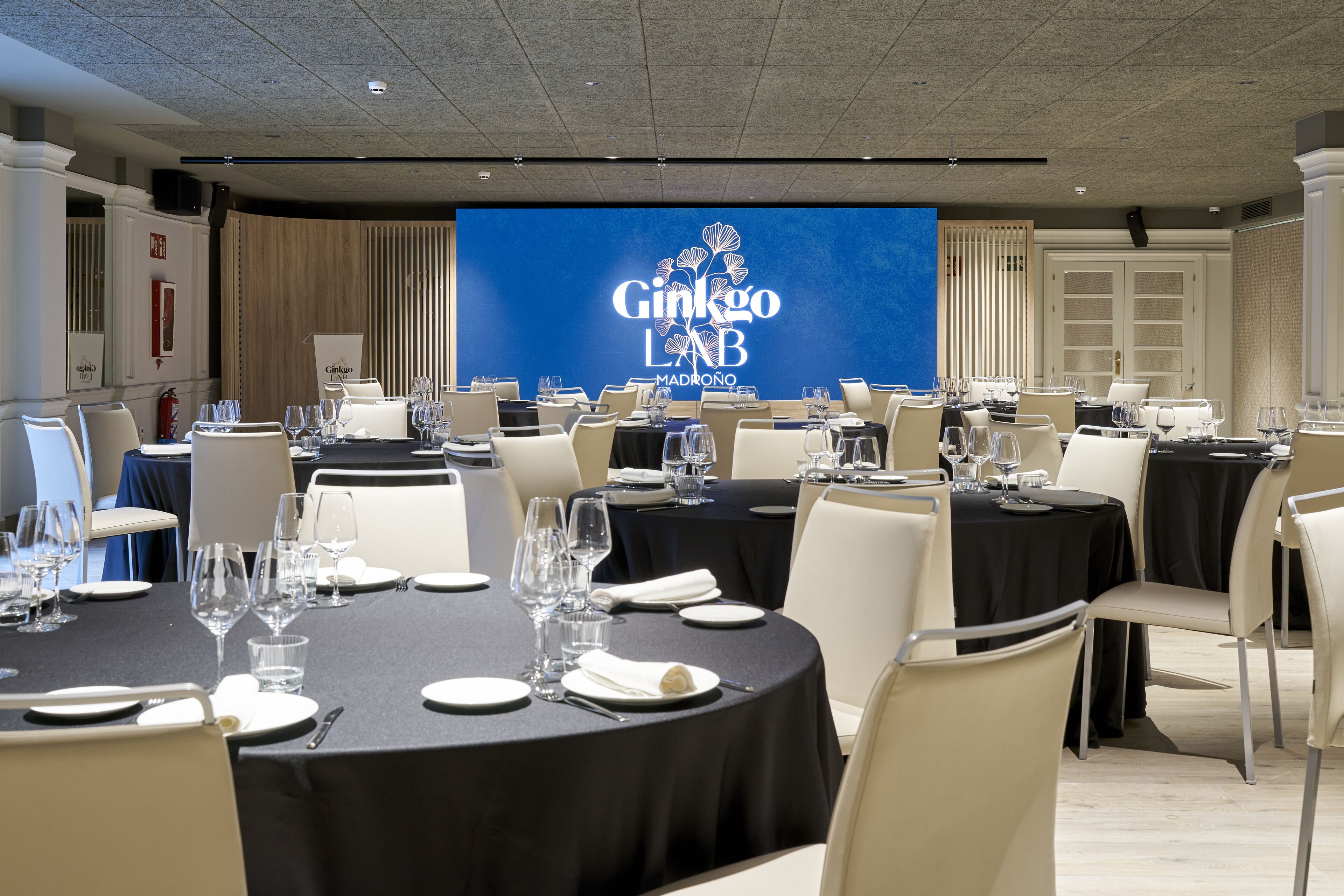 A conference room with round tables set for a meeting, a large screen displaying the Ginkgo LAB MADRÓNIO logo, and a modern interior design.