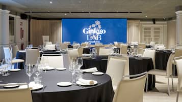 A conference room with round tables set for a meeting, a large screen displaying the Ginkgo LAB MADRÓNIO logo, and a modern interior design.