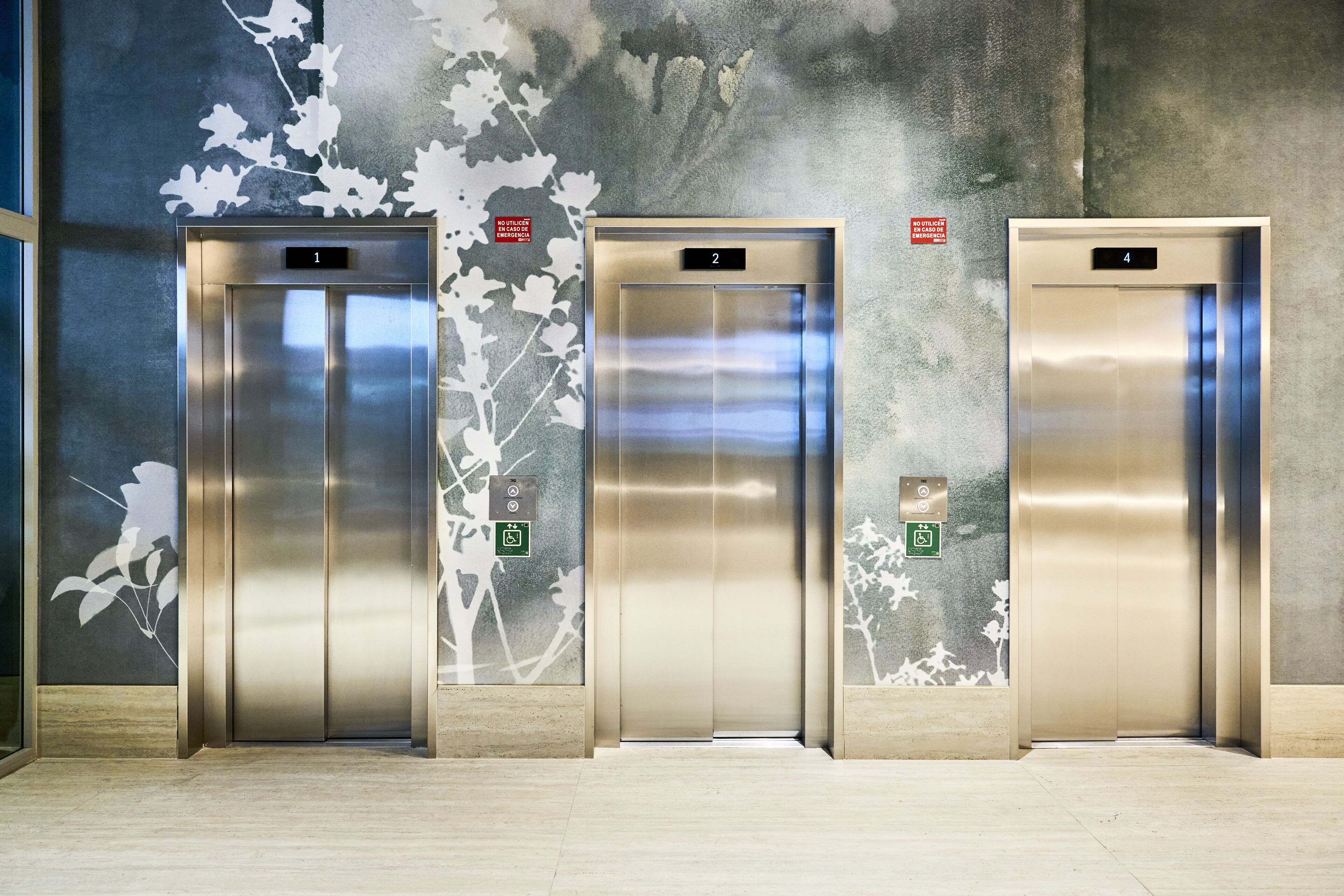 Three elevator doors with floor numbers 1, 2, and 4, and a wall with a leaf pattern and emergency exit sign.