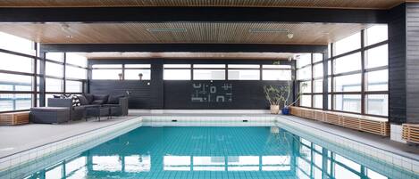 Una piscina interior moderna con pisos y paredes de baldosas, techo de madera y ventanas amplias.