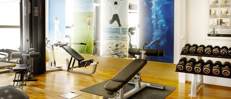 Un gimnasio con equipo de ejercicio, una banca, una pared con carteles de fitness y un estante con mancuernas.