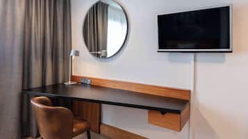 A modern hotel room with a wooden desk, a round mirror, a flat-screen TV, a chair, and a lamp.