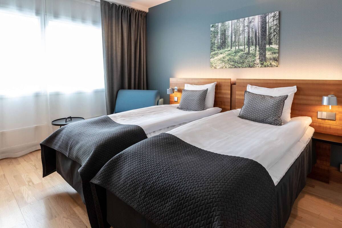 Hotellihuone, jossa on kaksi sänkyä, työpöytä, tuoli, taulu ja ikkuna, jossa on verhot.