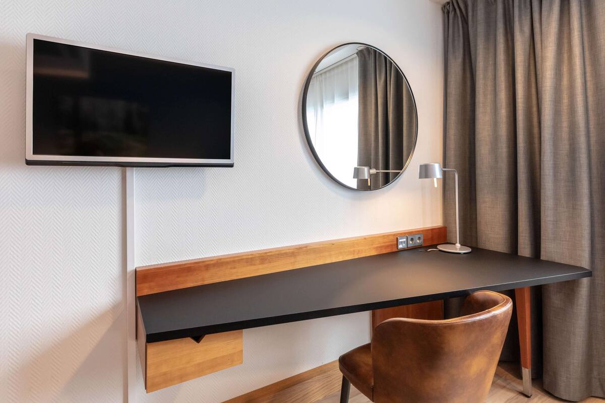 Moderni hotellihuone, jossa on seinälle asennettu taulutelevisio, pyöreä peili, lamppuvarustettu työpöytä ja nahkainen tuoli.