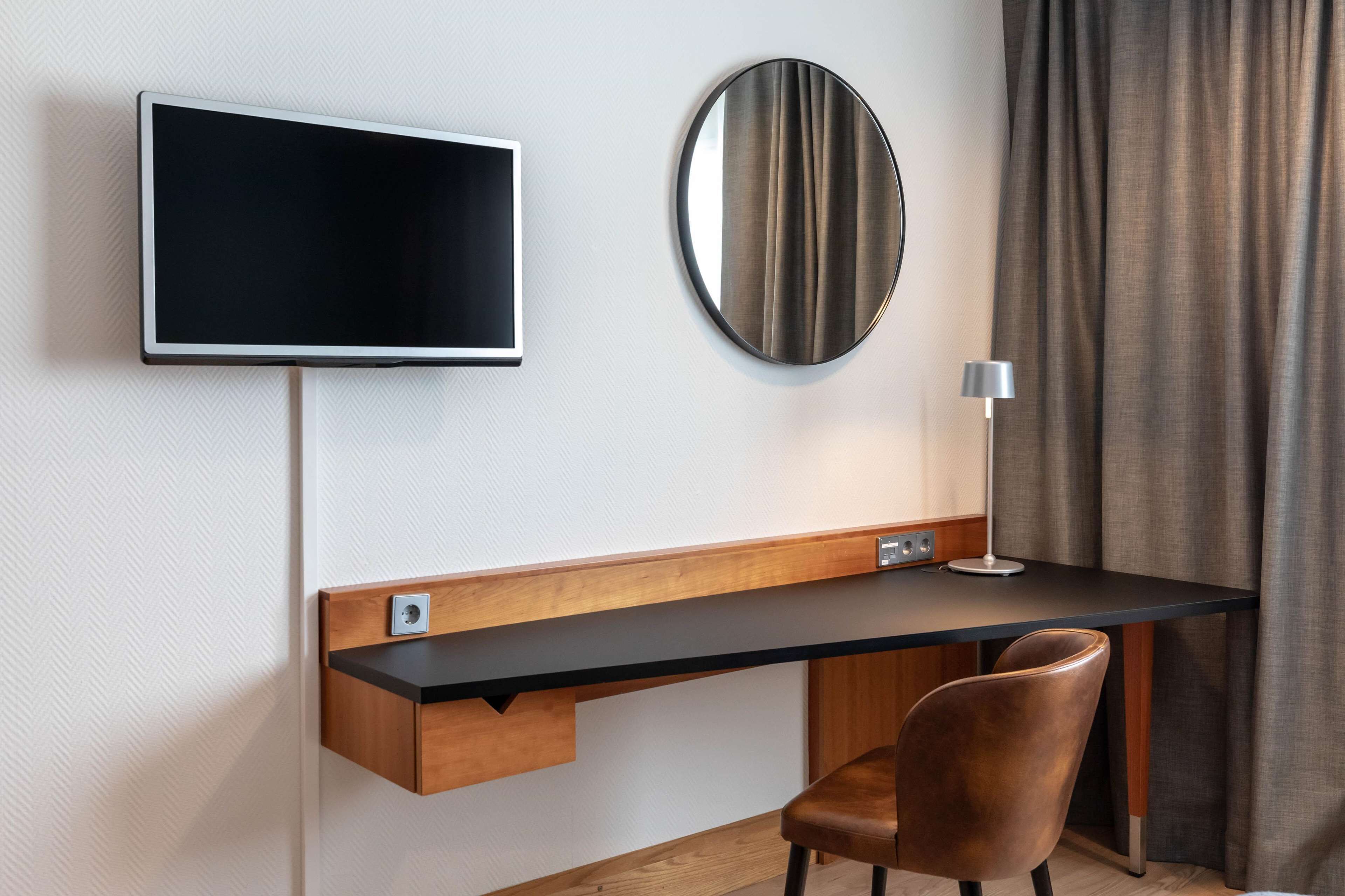 Una habitación de hotel moderna con un televisor de pantalla plana montado en la pared, un espejo redondo, un escritorio con una silla y una lámpara.
