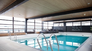 An indoor swimming pool with clear blue water, surrounded by a tiled floor and wooden ceiling. There are metal pool ladders, a seating area with sofas and a coffee table, and large windows offering a view of the outside. - Scandic Espoo (Espoo)