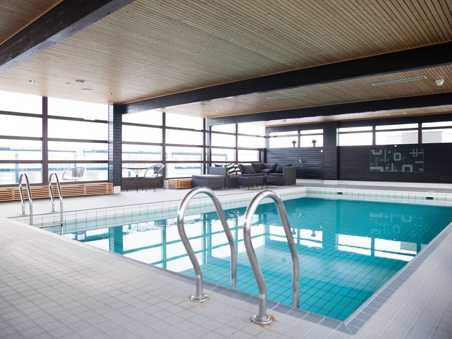 Una piscina interna con acqua cristallina, circondata da un pavimento piastrellato e un soffitto in legno. Si vedono scalette metalliche, un’area relax con divani e un tavolino da caffè, e ampie finestre che offrono una vista sull'esterno.