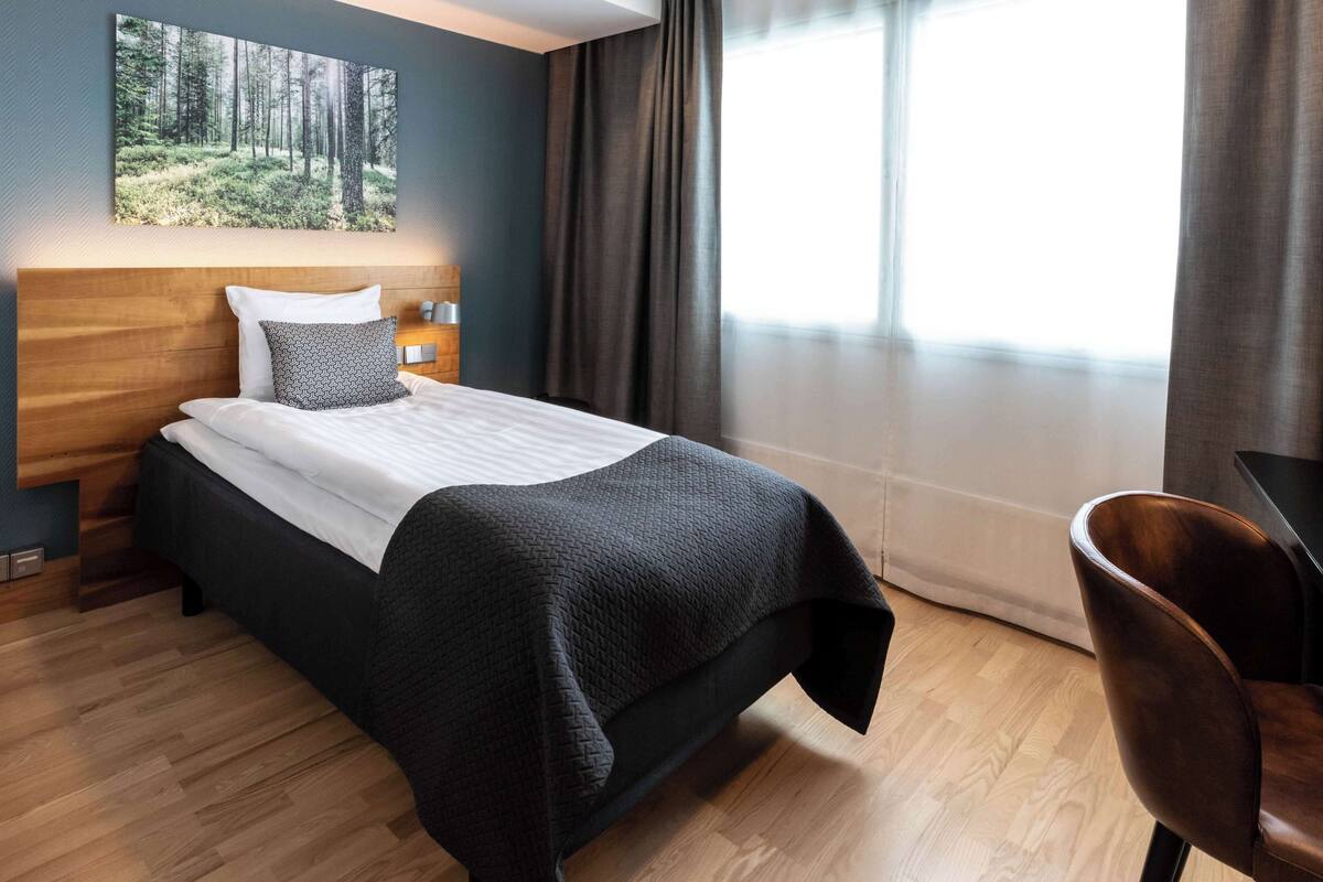 Hotellihuone, jossa on sänky, puinen pääty, yöpöytä, työpöytä ja suuri ikkuna, jossa on verhot.
