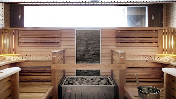 A sauna with wooden benches, a central stone wall, and a metal bucket on the floor. - Scandic Espoo (Espoo)