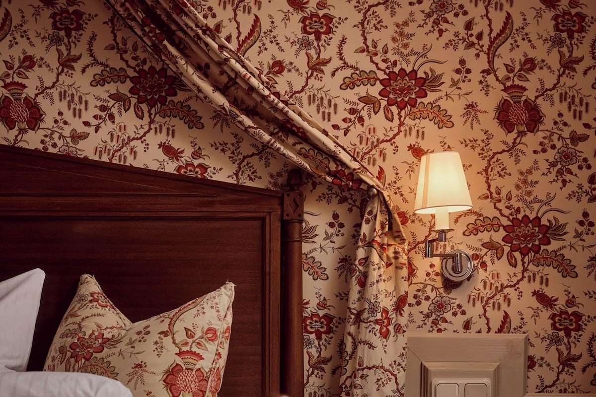 A bedroom with a bed, a nightstand, a lamp, and floral wallpaper.