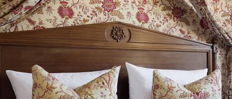 Un lit avec du linge de lit blanc et des oreillers à motifs floraux, une tête de lit en bois et un rideau à motifs floraux.