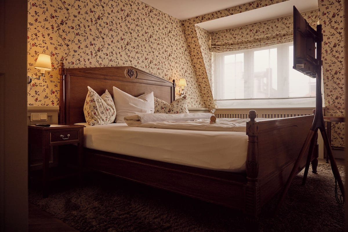 A neatly made bed with a wooden headboard, two bedside lamps, a small table, and a window with a view.
