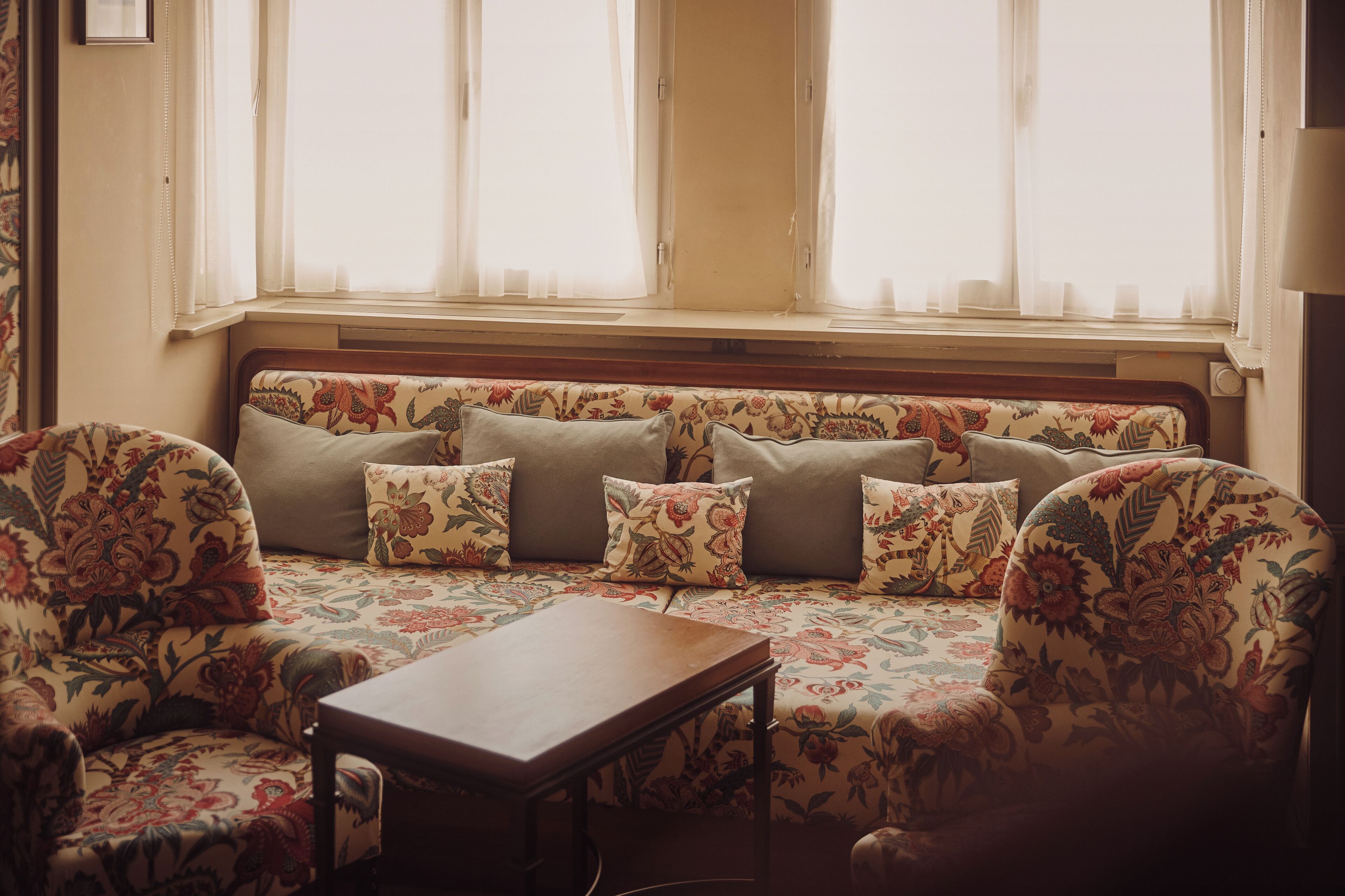 Una habitación con un sofá estampado con motivos florales, una mesa de centro de madera y una ventana con cortinas ligeras.