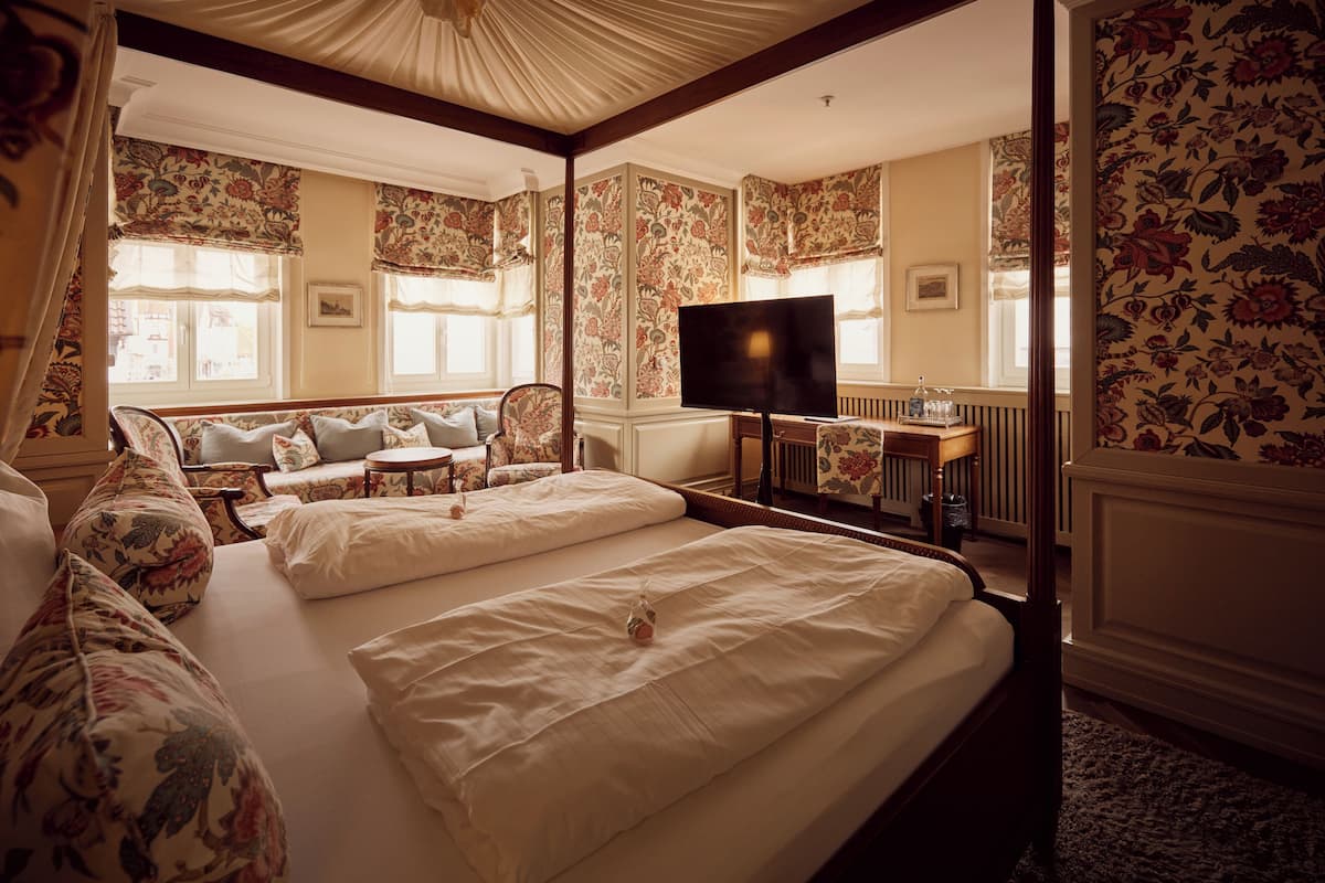 A hotel room with two beds, a television, a small table, and a window with floral wallpaper.