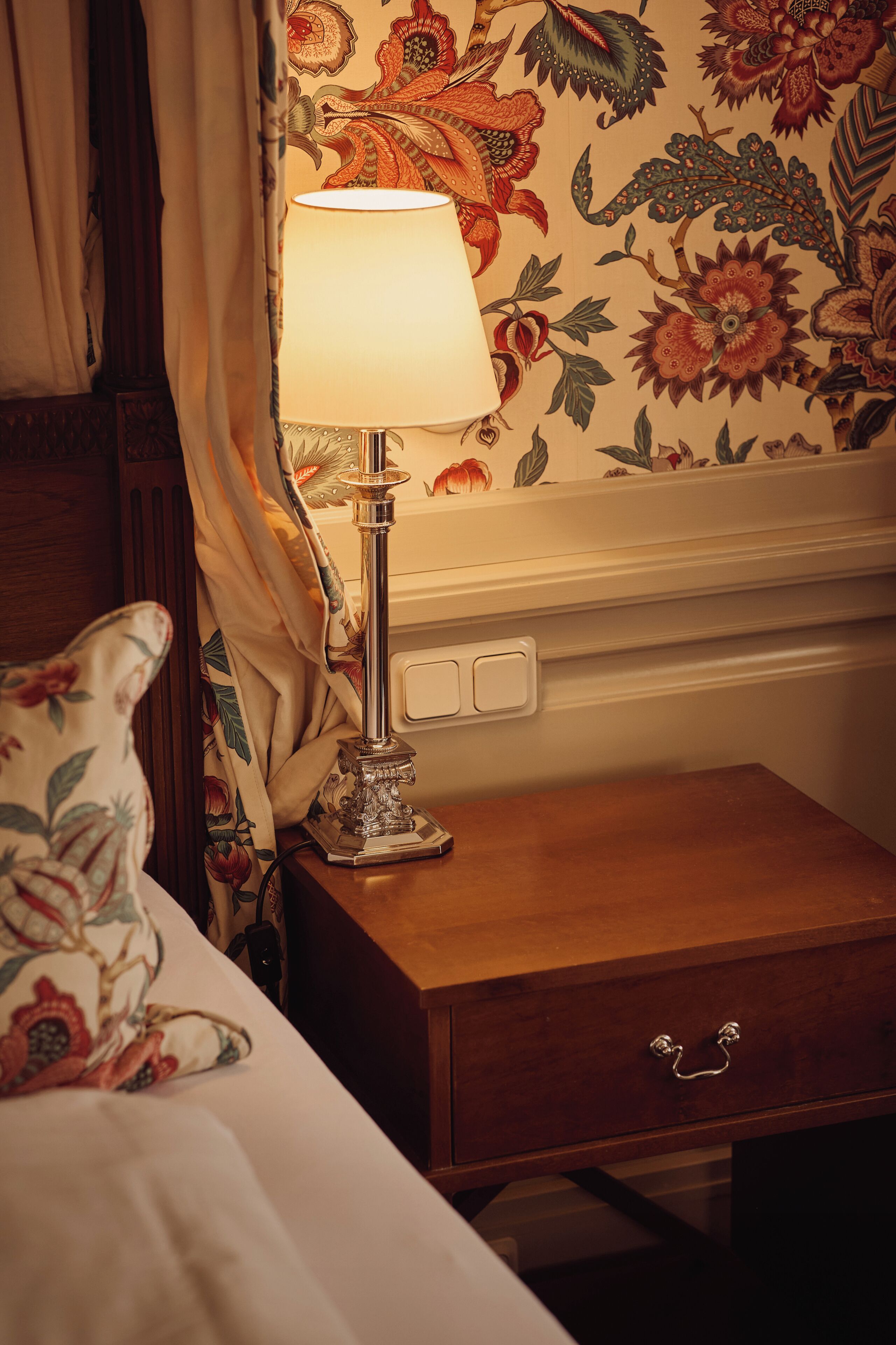 Un lit avec un oreiller à motifs floraux, une lampe de chevet, une table de chevet en bois et un mur recouvert d’un papier peint à fleurs.