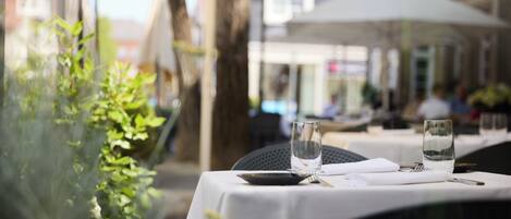 Un espacio de comedor al aire libre con mesas dispuestas para dos, cristalería transparente y vistas a la calle.