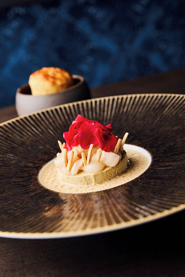 Un dessert servi dans une assiette sombre à bord texturé, agrémenté d’une quenelle de crème et d’une fleur rouge en guise de décoration.