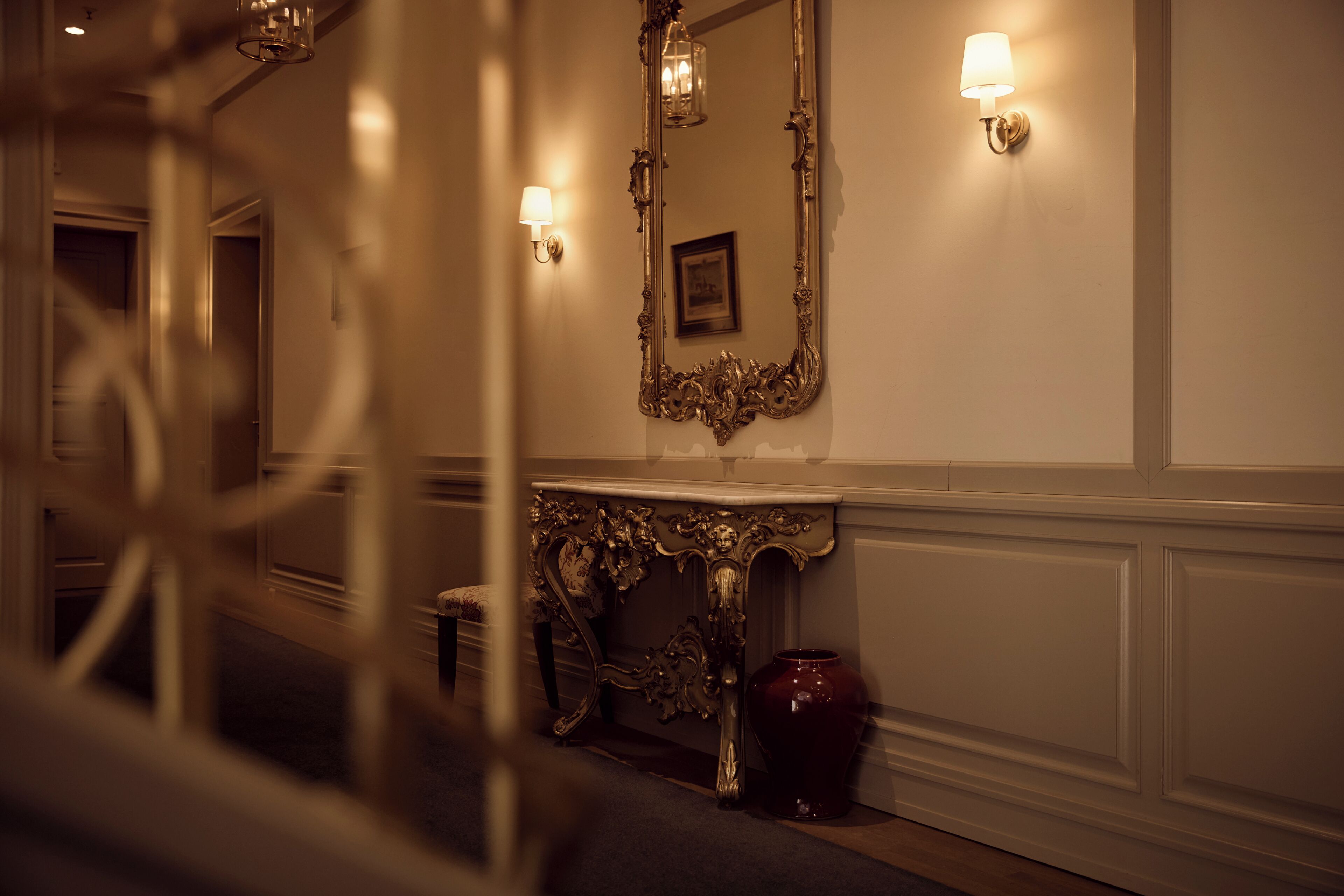 A hallway with a mirror, wall sconces, a console table, and a vase.