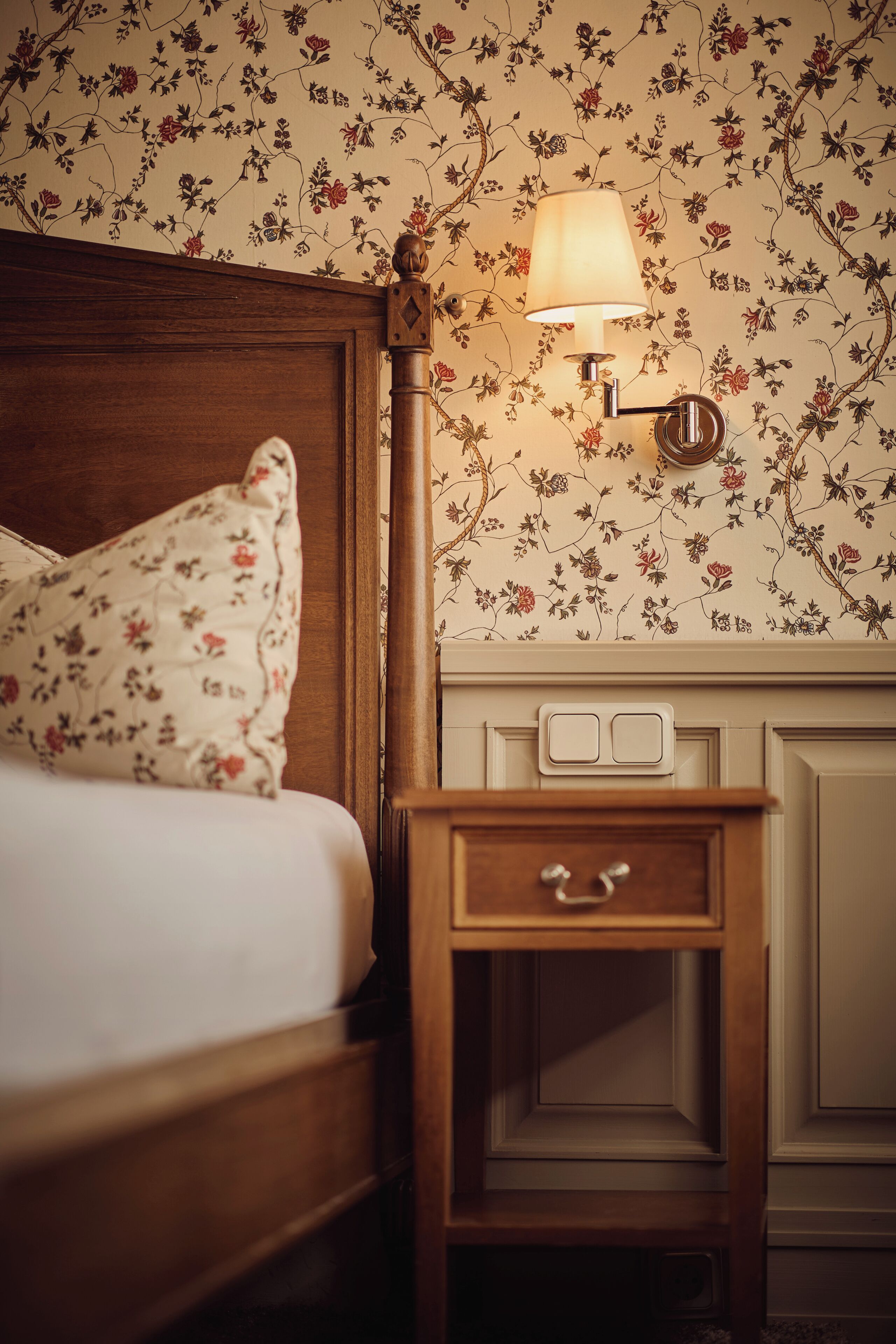 Un dormitorio con cabecera de madera, almohada estampada con motivos florales, mesita de noche con lámpara y papel tapiz floral en la pared.