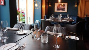 Une table à manger est dressée avec des verres à vin, des assiettes et des couverts. En arrière-plan, on aperçoit un mur bleu et un tableau encadré.