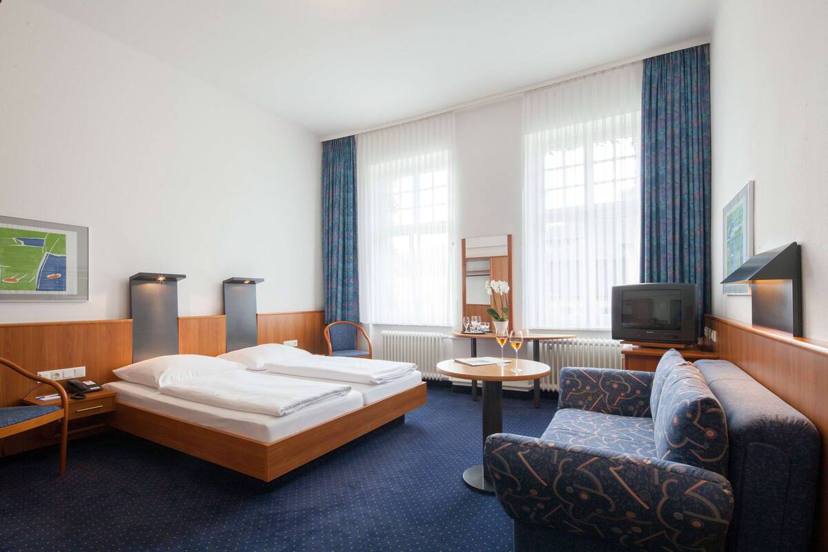 Habitación de hotel con cama, sofá, televisión, una mesita con un florero y copas de vino, y una silla.