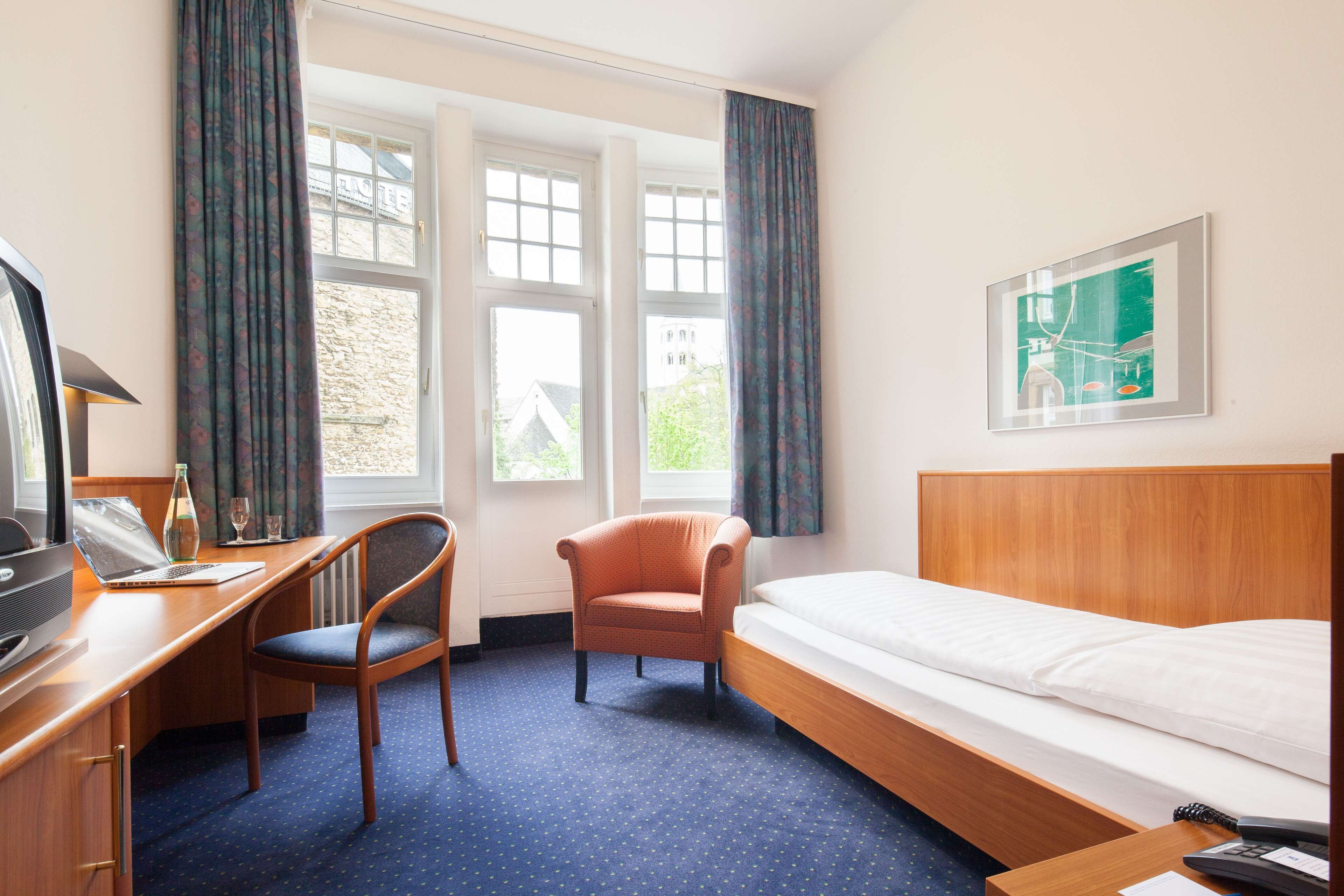 A hotel room with a wooden desk, a chair, a bed, and a window with curtains.