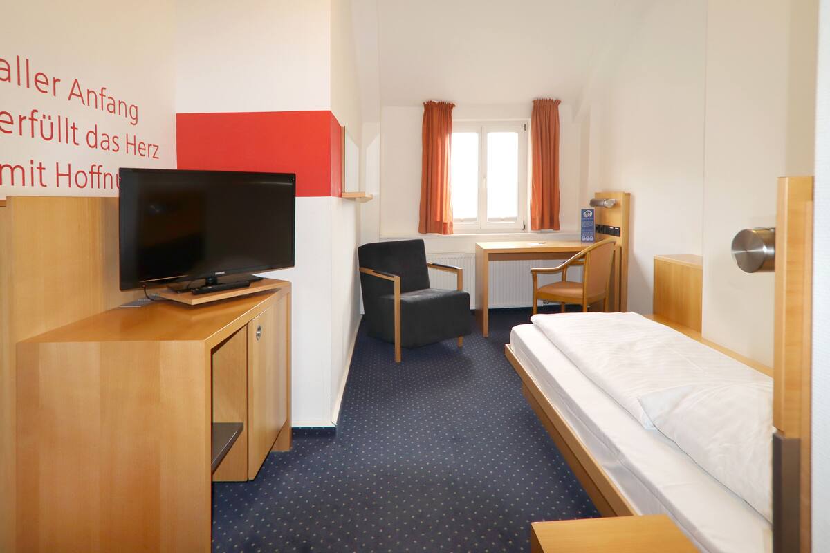 Habitación de hotel con cama, escritorio, silla, televisor y ventana con cortinas.