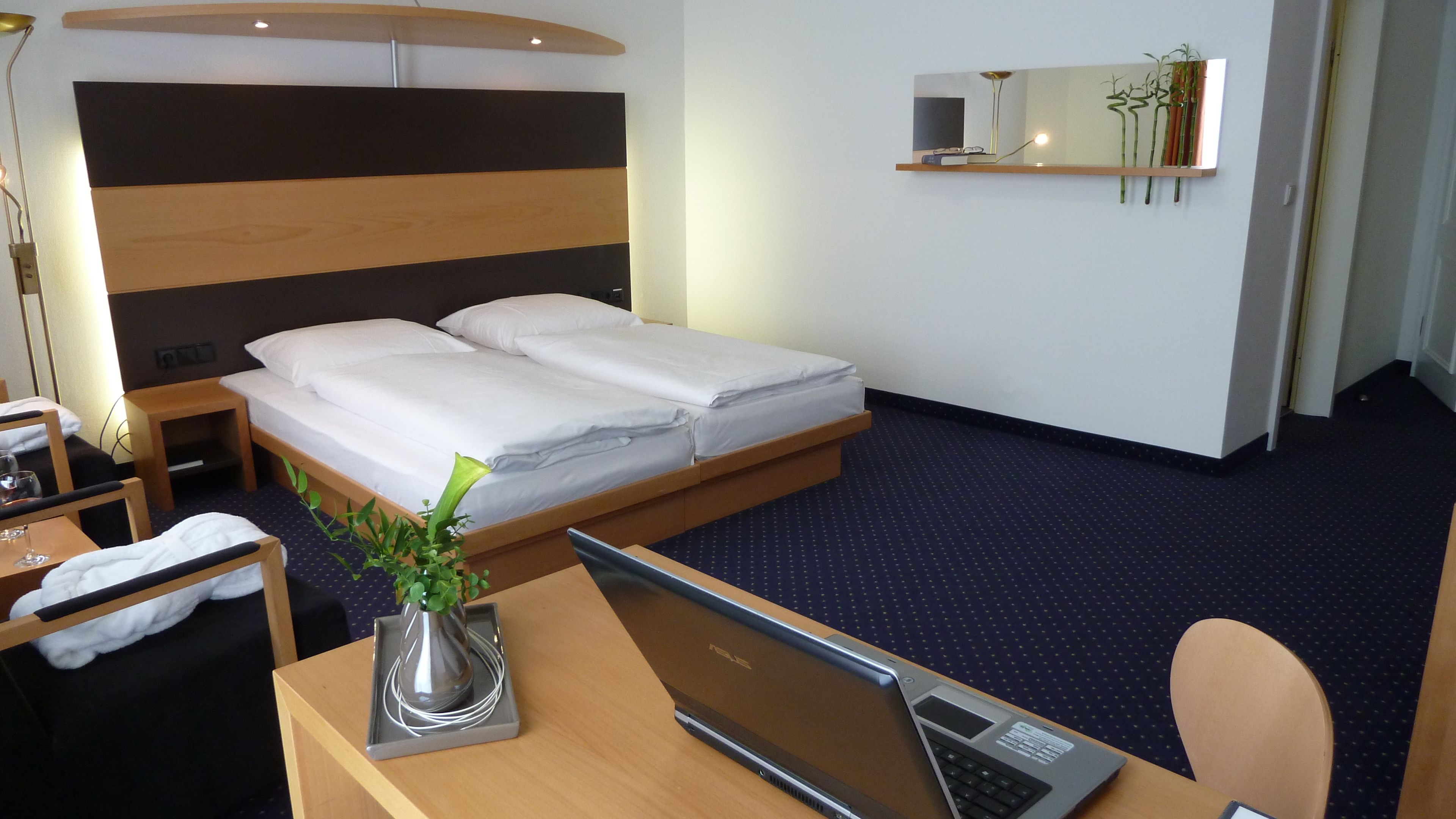 Ein Hotelzimmer mit Bett, Schreibtisch mit Laptop, Stuhl, kleinem Tisch mit Pflanze und Nachttisch.