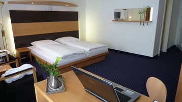 Quarto de hotel com cama, escrivaninha com um laptop, cadeira, pequena mesa com uma planta e mesa de cabeceira.