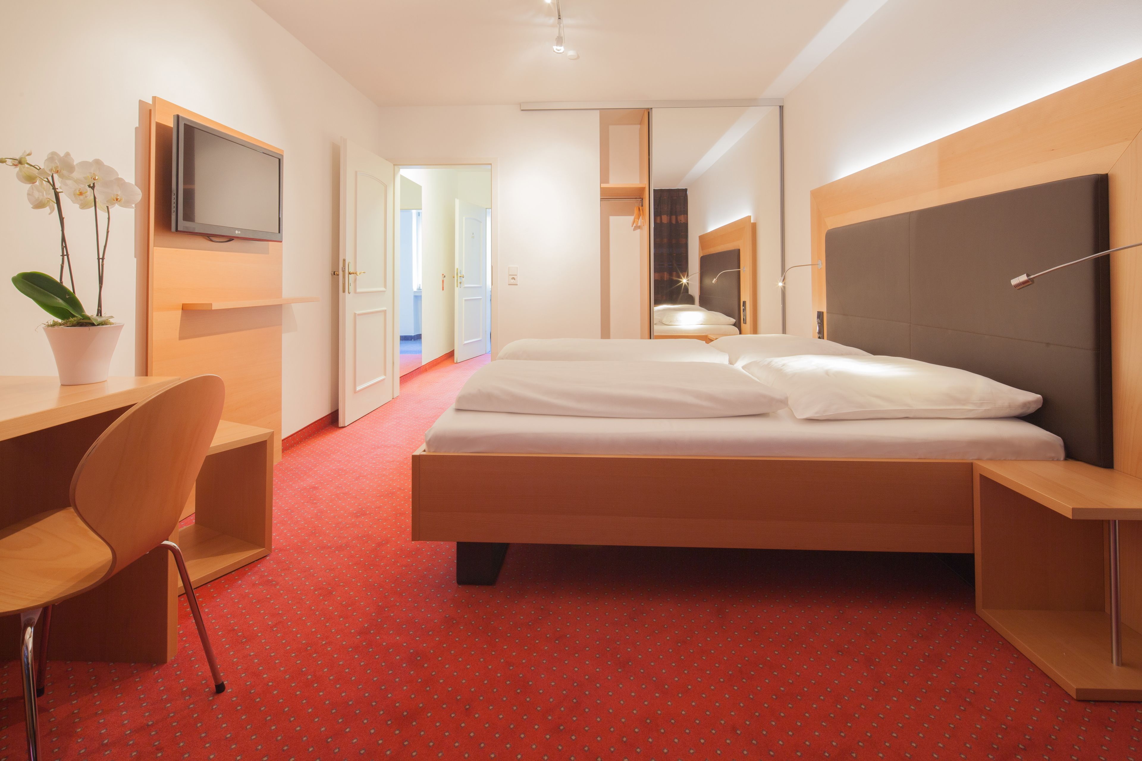 A modern hotel room with a large bed, a desk with a chair, a TV, and a potted plant.
