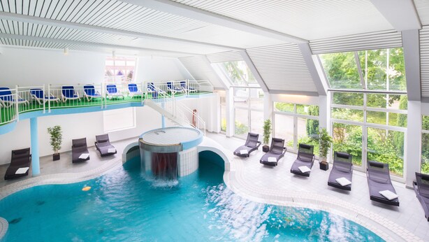 An indoor swimming pool with a central waterfall feature, lounge chairs, and a sun terrace with a view of greenery.